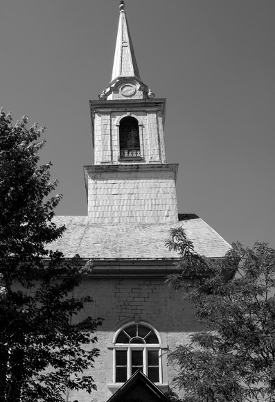 Black and white photograph of a pointy and leaning church bell tower taken from below and from one side of the church. Under the tower is a window and just below, the top of a side entrance door can be seen. Two trees are growing on each side.