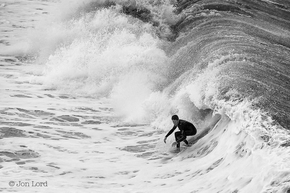 This is a black and white photo of a surfer surfing in front of a large wave.
Stretching upwards from the lower right corner to the upper right is a large and angry breaking wave. Below and to the right of centre is a young male teen surfer. He is wearing a black wetsuit, viewed in profile and looking towards, but not at the camera. He is moving diagonally from right to left and towards the camera. He is crouching forwards with his arms outstretched, his feet are spread, with one towards the back and one to the front of the white surfboard. Behind our surfer is the curling wave about to break, no more than a metre away. Beyond, the wave has already broken creating a maelstrom of white surf. Behind the wave, the water is dark, in front its a mix of black and white.
The location: Hermosa Beach, California (2014).

