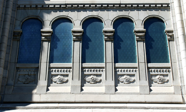 Photograph of a series of five arched windows on the side of a grey stone church, with opaque blue glass and a flower engraved under each one.