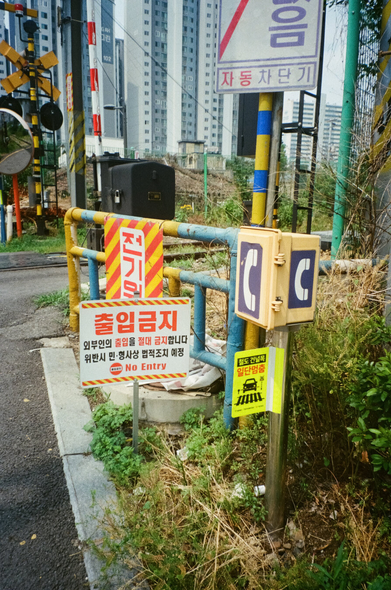 건널목 앞 구조물. 노란색·빨간색 등이 들어간 출입금지, 전기위험, 일단멈춤, 자동차단기 안내판과 긴급전화 상자 등이 배치되어 있고, 주변에 잡초가 많이 자랐다.