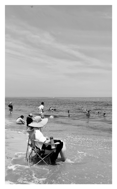 Monochrome photo of a scene at Sandy Hook Beach, New Jersey. The shore is at left and the ocean is at right, with waves rolling in. People sit or stand in the shallow part or wade deeper into the water. Prominently in the foreground, an older woman sits in a beach chair facing the sea, as the waves wash over her feet and the base of the chair. A water bottle sits in the holder of the armrest. The woman wears a white top, a black skirt pulled back to expose her calves, and a floppy sun hat that hides her face. Her reclining pose is very reminiscent of James McNeill Whistler’s famous portrait of his mother, although she’s facing the opposite direction. The sky above is streaked with high clouds, and a small dot of an airplane can be seen at the photo’s upper edge.