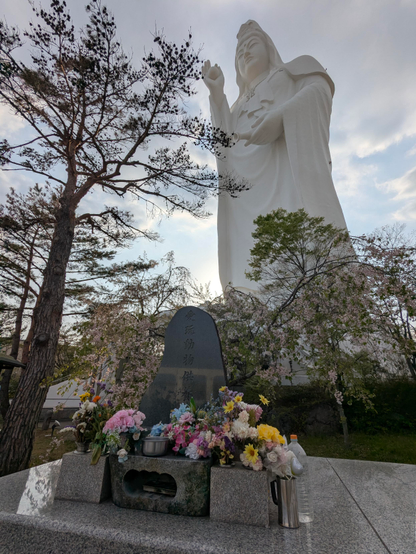 "Die Statue stellt den Bodhisattva Kannon aus dem Shingon-Buddhismus dar. Die weiße Frauenfigur hält in der rechten Hand eine Kugel mit einem Durchmesser von 3 m, die den Nyoihōju (如意 宝珠), einen Edelstein, darstellen soll, der seinem Besitzer Wünsche erfüllt. In der linken Hand hält die Figur eine Flasche (Länge 8 m und Durchmesser 2 m), die das Wasser der Weisheit enthalten soll.# (laut Wikipedia)

Die Statue wurde aus der Nähe und im Gegenlicht aufgenommen.
Daher scheint sie perspektivisch etwas nach links zu kippen.
Im Vordergrund ein Blumen Arrangement auf einem marmorierten Sockel und ein Baum, der ins Bild ragt.