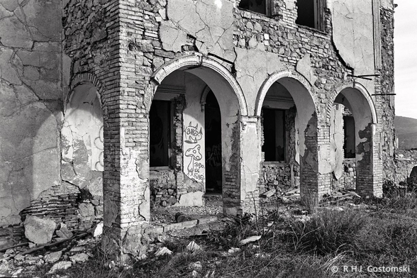 A black and white film photo of the front facade of an abandoned cortijo, featuring three arches. Most of the render has spalled exposing a mixed brick and stone construction