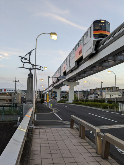 Die aufgeständerte Einschienenbahn mit silber-orangen Wagen fährt an mir gleich rechts vorbei.
Ich befinde mich auf einem Fußgängerweg über eine Brücke.
Bogenlampen haben sich bereits in der Dämmerung eingeschaltet. 