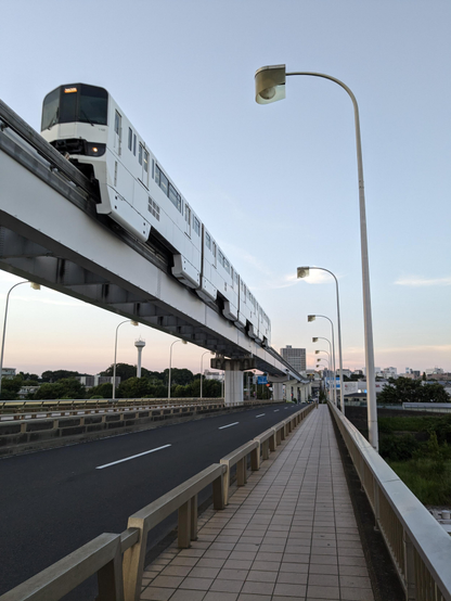 Die aufgeständerte Einschienenbahn mit weissen Wagen fährt an mir gleich links vorbei.
Ich befinde mich auf einem Fußgängerweg über eine Brücke.
Bogenlampen haben sich bereits in der Dämmerung eingeschaltet. 