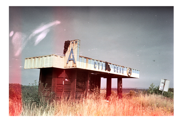 An abandoned bus stop stands overgrown with tall grass, its faded sign reading ‘Старе село’ partially obscured. The structure’s paint is peeling, and a tilted road sign leans nearby. The film shows light leaks, adding to the sense of decay and forgotten time.