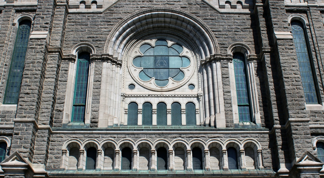 Photograph of a gothic-style church facade featuring a large, decorative rose window with a floral pattern and multiple arched stained glass windows. The building is constructed with grey stone, showcasing intricate ornate stonework and arches.