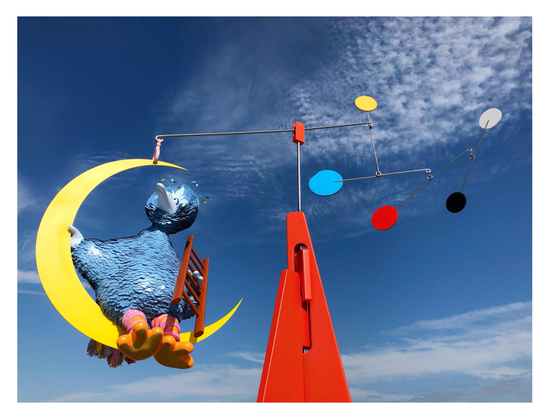 A wider view of the sculpture, viewed from a low angle, where we can now see Big Bird’s crescent moon suspended from one arm of a mobile attached to an orange triangular base. Multicolored circles are attached to other rotating arms. The sky beyond is blue with high cirrus clouds.
