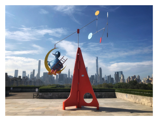 Another view of the sculpture, seen here on the Metropolitan Museum’s tiled rooftop bordered by the green hedges of its garden. Big Bird is suspended to the left of the sculpture’s base, looking off into the distance. Skyscrapers can be seen in the background under a blue sky with clouds.