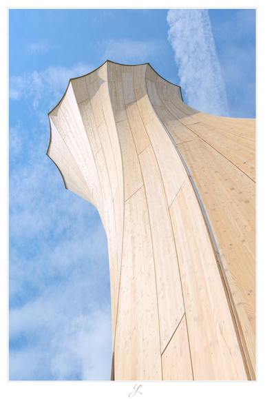 View of the blue sky along the outer wall of a wooden tower. The tower itself has a circular cross-section divided into approximately twelve sections, the edges of which are curved inward, giving the overall shape of the cross-section the appearance of a gently curved gear wheel. The wooden structure is twisted so that the cross-section twists counterclockwise by about 90° from the base to the top of the tower. The wood is light brown, the sky is light blue with a few white clouds and a dissipating contrail.

AI disclaimer: Using my work, its meta data, written or derived description to create media with or train AI based systems is prohibited.
