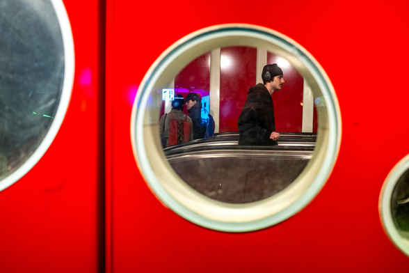 A red wall with small round glass windows of different sizes. A man dressed in black with a back wollen hat and a music headseat is seen through a window in the middle of the image. He is walking up an elevator.
The photo was taken at the De Brouckere metro station in Brussels.