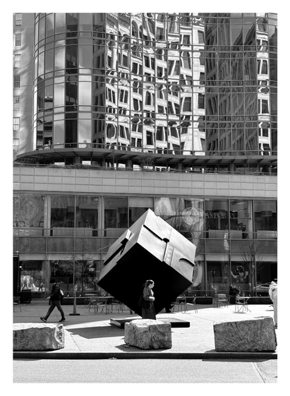 Monochrome photo of Tony Rosenthal’s sculpture “Alamo” (also known as The Cube or the Astor Place Cube), a large rotating black cube with recessed and protruding elements, mounted on one of its corners in the middle of a plaza in downtown Manhattan on a sunny day. Sunlight illuminates its upward-facing surfaces while casting its underside into shadow. Among a few pedestrians in the plaza, a woman in a coat stands out as she walks directly in front of the Cube and is framed by its black surface. In the foreground are three irregular concrete blocks, spaced out as traffic barriers at the edge of the sidewalk. Behind the Cube is some outdoor seating for a cafe, above which rises the Astor Place Tower. Its curving, wavelike facade of glass panels reflects the windows of other buildings but breaks them up into distorted rectangular fragments, reminiscent of Cubist paintings.