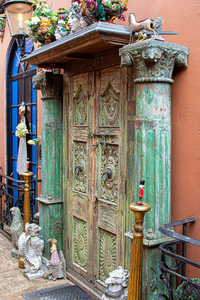 A double, wooden, green-stained door with six panels, all decorated. Photographed from the side. It's a combination of kitsch and art. Next to the door on either side are two large green pillars, and in front of them are two tall, wooden candlesticks topped with two tiny red toy soldiers. Above the door is a shelf with a row of heads in coloured, flowered hats, a toy horse on the pillar, and at the foot of the both pillars are a kind of stone devils and a small statue of a lady in a long skirt. Next to the door is an old-fashioned street lamp, and there's much, much more. The wall is orange.