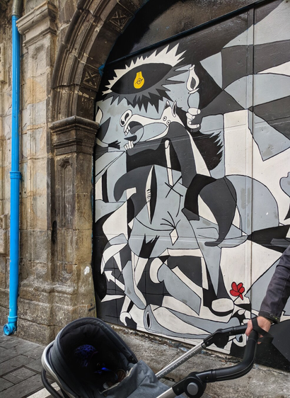 A street photograph of a colourful mural in the style of Pablo Picasso's Guernica, painted on a large black door inside a stone archway. The painting, in shades of grey, black and white, depicts a frantic, stylised scene with distorted figures, including a horse, a bull, and a person holding a candle. Above the scene, a lightbulb with a yellow filament hangs from a jagged sun-like shape. A single red flower, a detail not in Guernica, is painted near the bottom right. To the left, a bright blue drainpipe runs vertically down the stone archway. In the foreground, the handle and top of a dark grey baby buggy is visible, pushed from the right by a person wearing a black jacket. The ground is paved with grey and tan blocks.
