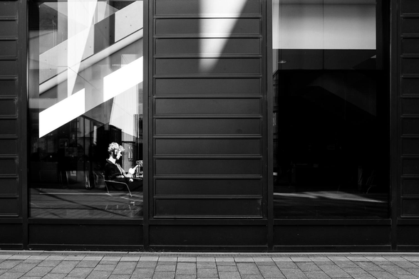 The image captures a solitary individual, engrossed in reading while seated indoors. The scene is framed by a large window, reflecting the external architecture and casting shadows that interplay with the interior space. 