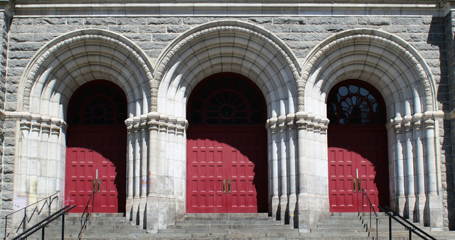 Photograph of the main entrance of a grey stone church, showing three red doors on top of stairs, in Gothic style with very ornate arches engraved in stone and semi circular window on top of each.