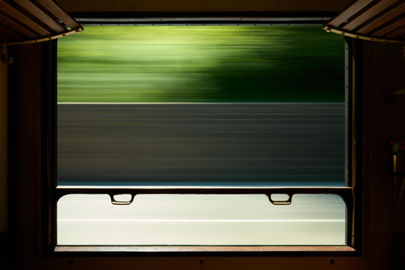 The image shows a view from a train window, with blurred green foliage and a gray background indicating rapid movement. The window frame is partially lit, highlighting the stillness of the interior of the train compartment.