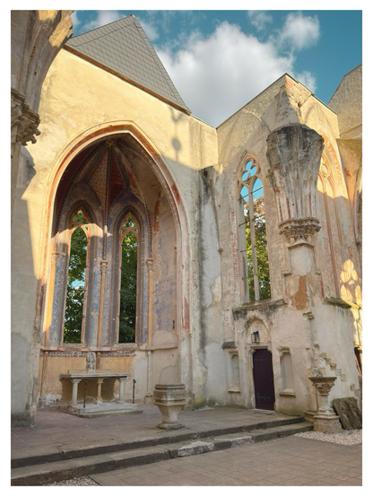 Interior view of a church ruin with tall Gothic arches, partially preserved frescoes, and windows without glass. The altar and stone structures remain in place, surrounded by weathered walls with faded decorative patterns. Sunlight highlights the worn stone, while trees are visible through the open windows under a partly cloudy sky.