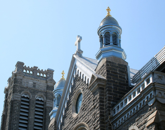 Photograph of the top parts of a gothic inspired church, with a grey stone square tower bell, two turrets with silver aluminum roof and cladding surmonted by fleur-de-lys spikes, and grey stone main building with aluminum roof, cross on top and parapets.