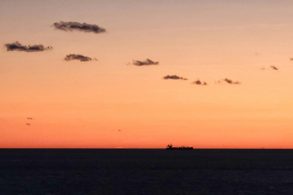 The black silhouette of a cargo ship against a very orange and yellow sky with a few small, puffy grey clouds.