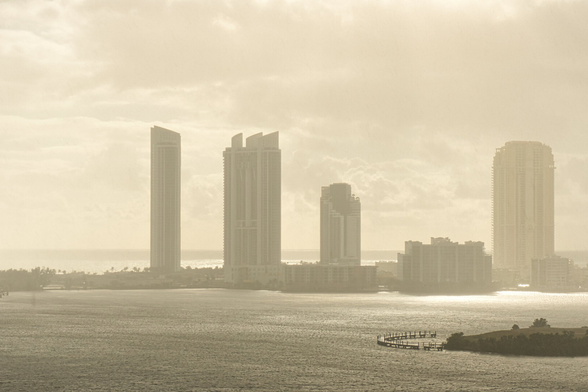 A city skyline with tall buildings silhouetted against a sky that is cloudy, sunny, and rainy. The scene is bathed in a soft sepia tone, creating a calm and nostalgic atmosphere.