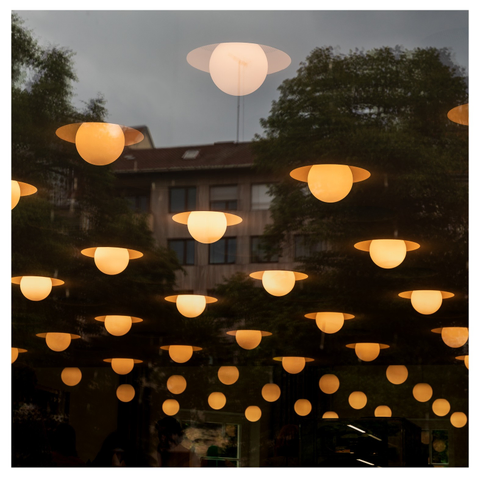 Foto einer Reflexion auf einer Fensterscheibe, die ein gegenüberliegendes Haus und Bäume zeigt. Durch das Fenster schimmern auch eine große Zahl von Deckenlampen des dahinter liegenden Raums. 