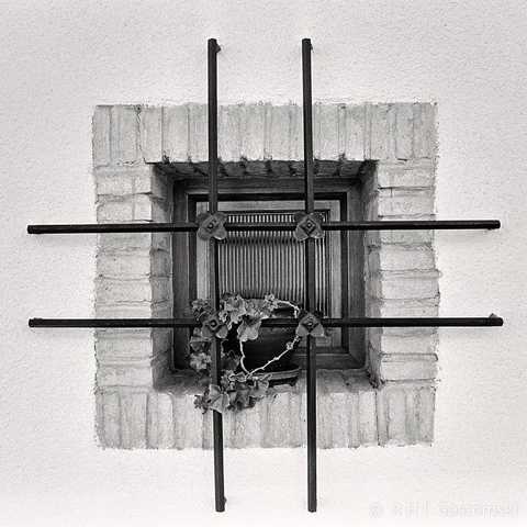 A small, square window with vertical and horizontal metal bars, set in a brick frame on a white wall in Segura de la Sierra. A potted plant with wilted leaves sits on the sill behind the bars, captured on Ilford FP4 film.