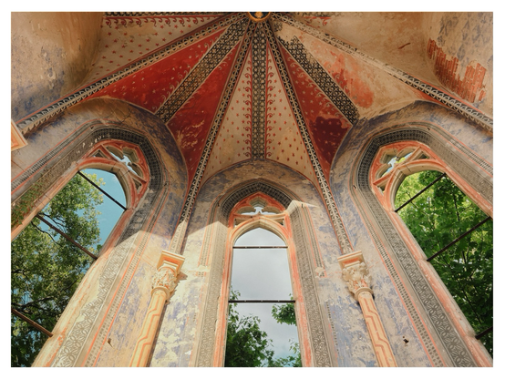 View from inside a church ruin looking up at tall Gothic arched windows without glass. The vaulted ceiling is decorated with faded red, black, and white painted patterns, and the walls show remnants of ornate frescoes. Through the open window frames, green trees and the sky are visible.