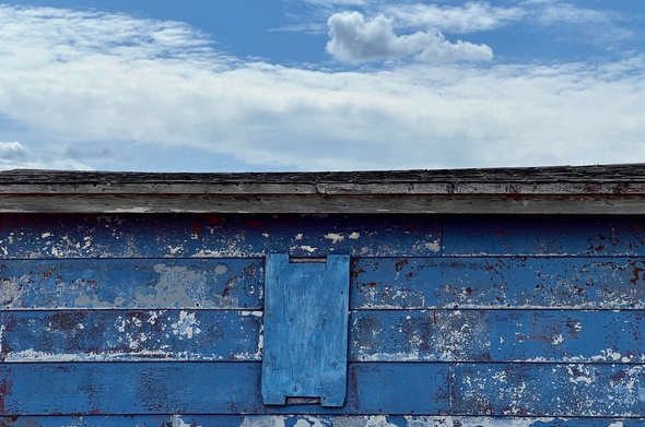 A boarded window near the roofline on a weatherbeaten wooden shed. The blue paint on the siding is peeling, and the board covering the window is stained a similar shade of bright blue. The black shingled roof is partially visible due to the camera angle. The sky’s blue is similar to the shed colour, with a thick band of wispy white clouds and one small  prominent cumulus cloud. [Cape Freels (NL, Canada) May 2025]