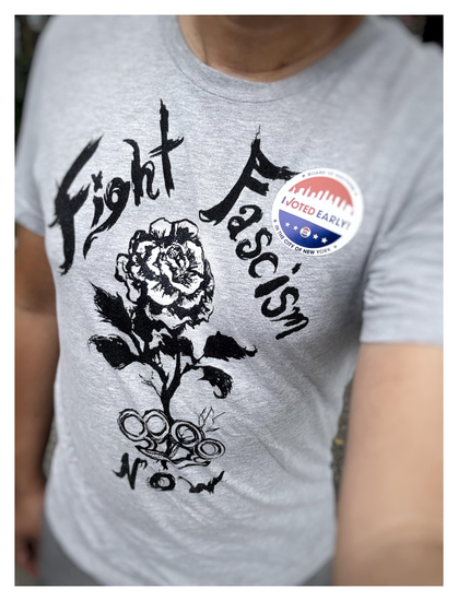A selfie of my torso wearing a gray T-shirt with an ink illustration of a rose blooming out of a set of brass knuckles, surrounded by the words “Fight Fascism Now.” On the left (viewer’s right) side of my chest is a circular red-and-blue “I Voted Early” sticker from the New York City primary elections.