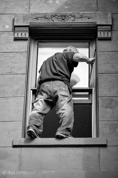 This is a black and white photo of a man cleaning a window.
In the centre of the image is a middle aged man standing on the stone window sill on the outside of a first floor (US-second floor) window. The building has a light grey rendered facade with large brick like shapes carved into the plaster. Our window cleaner is standing on the stone plinth like sill supporting a sash window with the lower sash raised. Above the window is a large lintel with a delicate floral pattern inlaid in the centre. The window cleaner has his back to the camera, is wearing jeans and a black tennis shirt.
The location: Boston, Massachusetts (2013).