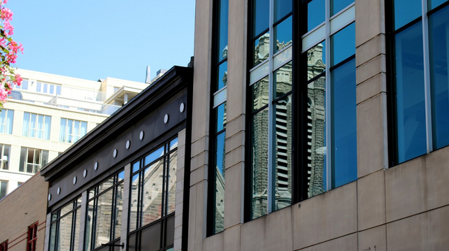 Photograph of rectangular windows of modern buildings reflecting a Gothic inspired church with a square tower bell on the other side of the street. Another white apartment building can be seen in the background, under a pale blue sky.