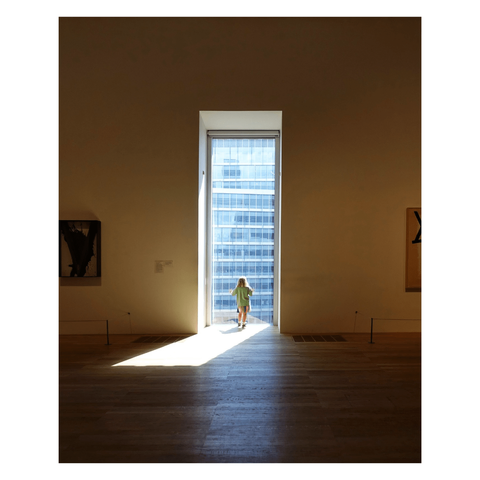 This is a portrait format colour image of a small child looking out of a large window in a modern art gallery. Taken from behind, the child stands in the middle distance with their hands against the glass. Light streams through the window, casting a long shaft of light across the otherwise dark wooden floor. Glimpses of the art displayed on the walls are just visible to the right and left of the frame. Taken with a Sony RX100 at the Tate Modern, London.