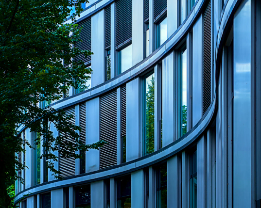 Drei Stockwerke voller Fenster schwingen sich in einer S-Form von rechts nach links durch das Bild. Die Fenster wechseln sich in jeder Reihe mit glatten Fassadenplatten ab. Die S-Form ergibt sich aus drei schmalen Streifen, die zwischen den Stockwerken entlanglaufen. 
Von links ragt ein dichter, grüner Baum in das Bild. Das ganze Gebäude macht einen kühlen, sehr modernen Eindruck. 
. 
Three floors full of windows swing in an S-shape from right to left across the photo. The windows alternate in each row with smooth front panels. The S-shape is made up of three narrow strips that run between the floors. A dense, green tree protrudes into the photo from the left. The whole building gives a cool, very modern impression.
.