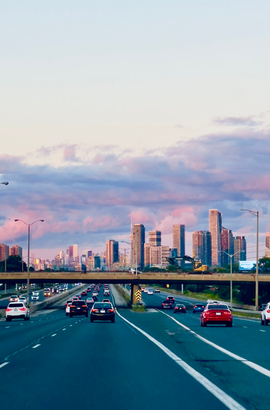 View from the highway towards downtown, a bridge crosses the highway and beyond that is the city, reflected pink and purple from the sunset.