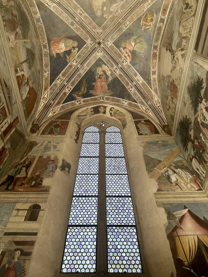 A tall window in a chapel wall, seen from the inside, no stained glass art, just lightly colored small hexagonal panels, seen from below, so both wall around it ad ceiling are visible. Wall and ceiling covered with elaborate and colorful frescoes telling various religious stories.