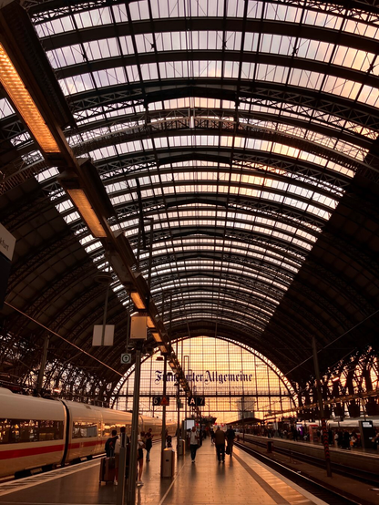 The photo shows the grand interior of Frankfurt’s main station at sunset. The vast iron-and-glass roof arches high overhead, its panels glowing in warm shades of orange and violet as the evening light filters through. At the far end, the huge glass façade shines brightly, carrying the bold lettering Frankfurter Allgemeine. Beneath, platforms stretch into the distance, lined with tracks that vanish toward the glowing horizon. On the left, a sleek white ICE train waits, its red stripe catching the light. Travelers move along the polished floor with luggage, some heading quickly, others strolling more slowly. Lamps and signs line the platforms, their artificial glow mixing with the natural radiance from outside. The scene blends historic architecture, modern travel, and the fleeting beauty of the setting sun.