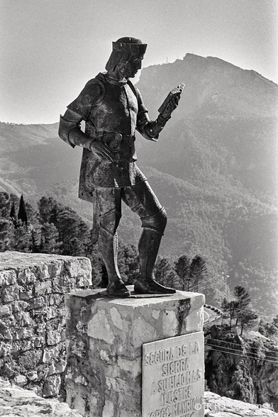 A statue of poet Jorge Manrique in historic clothing stands on a stone pedestal in Segura de la Sierra, overlooking a mountainous landscape with trees and a distant peak under a clear sky.