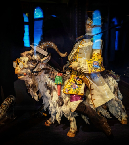 A colourful ceramic sculpture depicts a man on the back of a goat. The man, with a pale face and spectacles, is dressed in an ornate, old-fashioned coat with yellow and blue floral patterns, white frills, and a long white waistcoat. He wears dark brown trousers and has a serious expression. The goat has white fur with brown and black patches, and is also wearing spectacles on its face and forehead. Both figures are perched on a dark, reflective surface. The background is a deep blue and black, with the reflections of arched windows visible, suggesting the sculpture is in a display case. The overall style is intricate and fantastical.