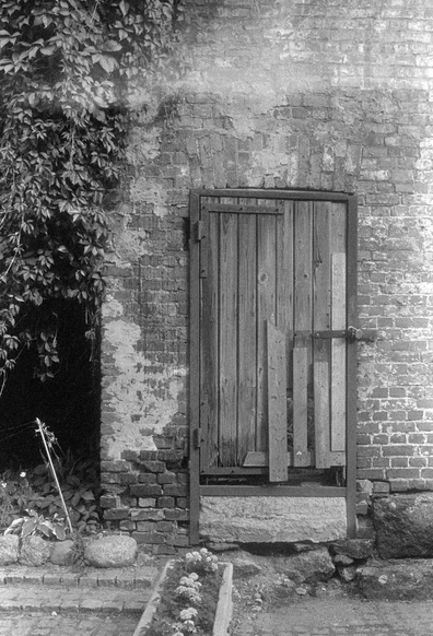 The black-and-white photo shows an old wooden door with a lock, built into a brick wall. The wall is dilapidated, with peeling plaster and exposed bricks. To the left of the door, there is a dense foliage. In front of the door, there are stone steps and a flower box filled with flowers.