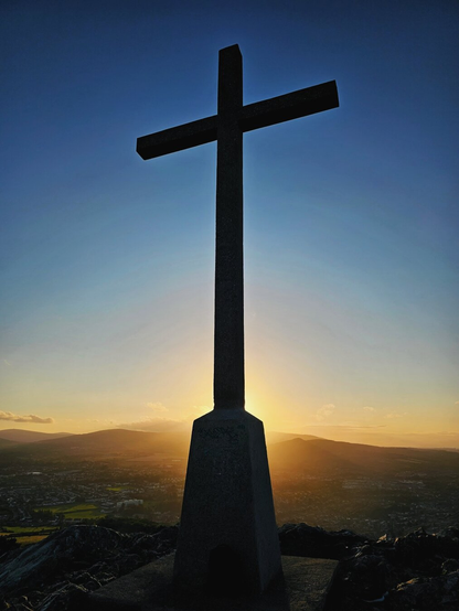 A silhouette of a large, stone cross standing atop Bray Head in County Wicklow, Ireland. The cross is positioned against a vibrant sunrise or sunset sky, with rays of light radiating behind it. The landscape below, including distant hills and a town, is visible but largely in shadow. The image has a calm, spiritual, and contemplative mood.