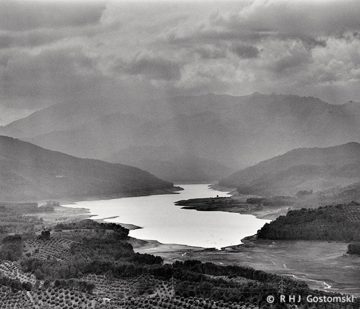 A black and white landscape photo, shot on Ilford FP4 with a Mamiya RB67, shows the Tranco reservoir surrounded by forested hills and mountains beneath a dramatic, cloudy sky. Dense trees and some open areas line the water’s edge.