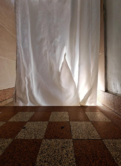 Interior photo of a bathroom with a white shower curtain between white walls. The floor features a checkered pattern of speckled brown stone tiles. Sunlight from an invisible window creates shadows on the floor through the translucent curtain fabric.
