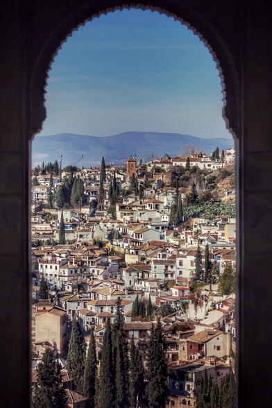 A stunning view of the cityscape of Granada, Spain, framed by a dark, ornate archway. The city is a dense collection of white buildings with terracotta roofs, nestled on a hillside. Cypress trees are scattered throughout the urban landscape. In the background, rolling hills and a mountain range are visible under a clear blue sky.