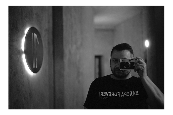 A black and white self-portrait taken in a mirror. A man holds a vintage rangefinder film camera up to his eye, wearing a dark T-shirt with reversed text visible in the reflection. The dim hallway is softly lit, with glowing wall lights framing the scene.