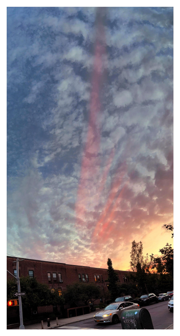 A Brooklyn residential street at sunset. Over low-rise apartments and light traffic, a red or monochrome rainbow — several bands of reddish light — swoops into a deep blue sky with dramatic clouds. The longest of the bands reaches past the top of the photo. The sun sets in a golden glow behind the silhouette of trees at lower right.