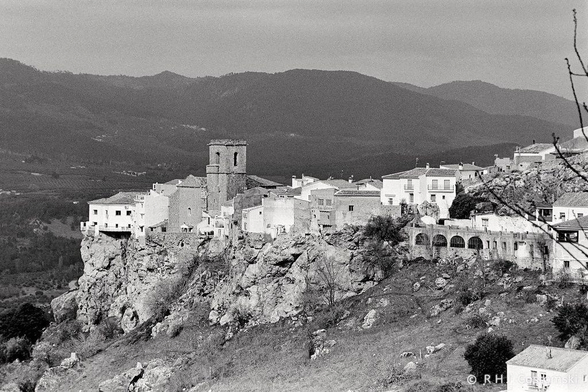 Black and white photo of a village with white buildings and a church tower perched on a rocky cliff, captured on Ilford FP4 film, set against a backdrop of mountains and forested hills.