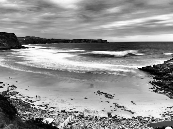Una fotografía en blanco y negro de un paisaje costero amplio. El cielo está cubierto de nubes dramáticas y en movimiento. Un mar oscuro se extiende hacia el horizonte, con olas difuminadas por una exposición prolongada que crean un efecto sedoso y etéreo al romper sobre la arena de la playa. A la izquierda y en el fondo, se ven acantilados rocosos y oscuros que bordean la costa. La playa, de arena clara, está salpicada de numerosas rocas de diversos tamaños, especialmente en el primer plano. Se aprecian algunas huellas y dos pequeñas siluetas de personas caminando cerca del agua en el centro de la playa. En la esquina inferior derecha, un camino o rampa con una barandilla desciende hacia la playa, rodeado de más rocas y vegetación oscura.