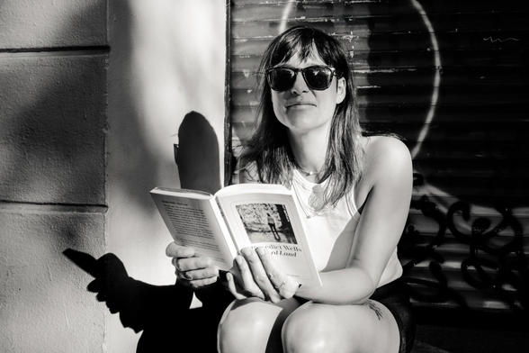 In this black and white photograph, a young woman sits in an urban setting. She is wearing sunglasses and is holding book. The background features a textured wall with her shadow on it on one side and a metal shutter with graffiti-like markings on the other.