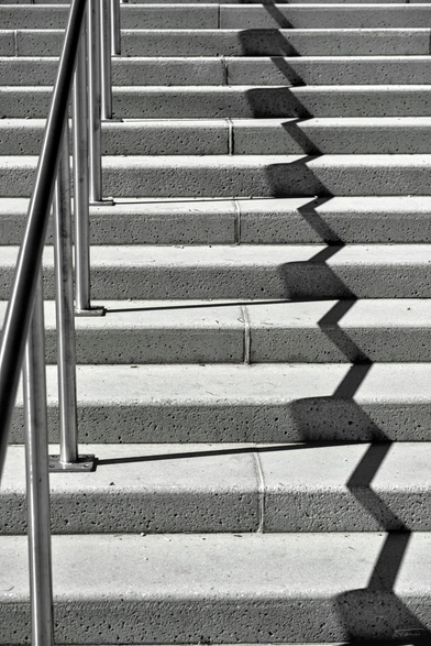 Das Foto zeigt eine Treppe in Schwarz-Weiß. Die Aufnahme ist aus einem ungewöhnlichen Winkel gemacht, der die Schatten der Geländer auf die Stufen wirft. Die Schatten bilden dabei ein interessantes, zickzackförmiges Muster auf den Stufen. Die Treppe selbst besteht aus Betonstufen und hat ein Metallgeländer auf der linken Seite. Das Spiel von Licht und Schatten verleiht dem Bild eine grafische und fast abstrakte Wirkung.